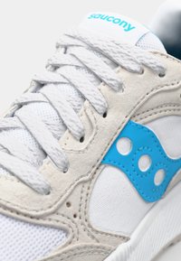 White sneaker with soft suede accents, blue rubber detailing, woven laces, and a textured mesh body. Features a distinctive brand logo.