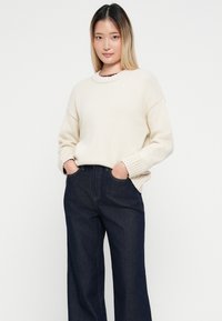 Strickpullover in Creme mit gerippten Bündchen und Rundhalsausschnitt, kombiniert mit dunkelblauen weiten Jeans. Das Model posiert mit den Händen in den Taschen.