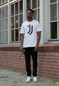 Hvid t-shirt med et sort logo, parret med sorte træningsbukser med hvide striber. Sneakers er hvide med en brun sål.