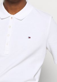 Vit polotröja i bomull med strukturfinish, med en treknäppt knäppning och en liten broderad logotyp i röd, vit och blå.