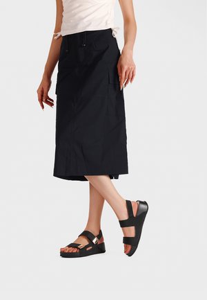 Donna che indossa una gonna midi nera con tasche laterali e sandali platform neri con due cinturini e dettagli metallici.