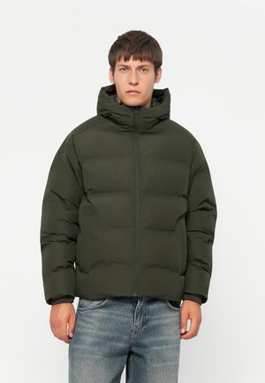 Man wearing dark green puffer jacket with hood and blue jeans, standing against plain white background.