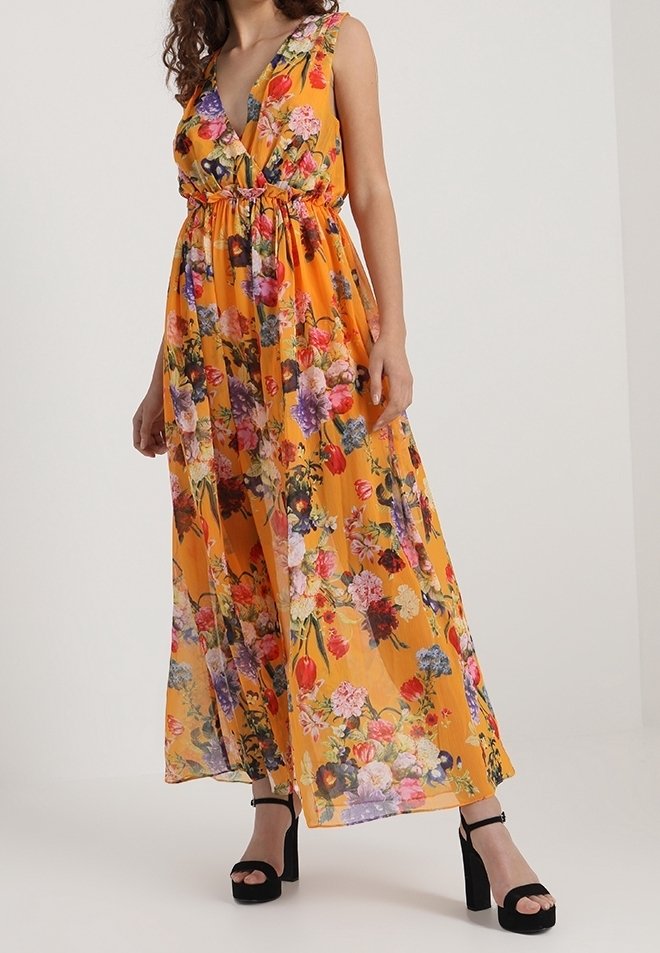Femme portant une robe maxi orange sans manches avec un imprimé floral multicolore et des sandales à talons hauts noires.