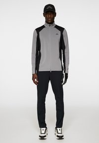 Szara marynarka sportowa z czarnymi akcentami, z wysokim kołnierzem i zamkiem błyskawicznym, zestawiona z czarnymi spodniami oraz białymi i czarnymi sneakerami.