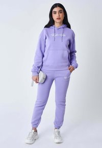 Felpa con cappuccio color viola chiaro e pantaloni da jogging abbinati realizzati in materiale morbido, con tasca frontale, coulisse e dettagli con logo. Abbinati a sneakers bianche.