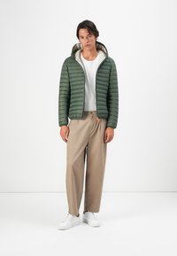 Tânăr îmbrăcat cu o jachetă verde matlasată deschisă, tricou alb, pantaloni largi bej și adidași albi, stând pe un fundal alb.