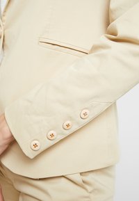 Veste blazer beige sur mesure mettant en valeur un tissu lisse, un revers net et six boutons ronds de couleur crème avec un motif croisé au niveau du poignet.
