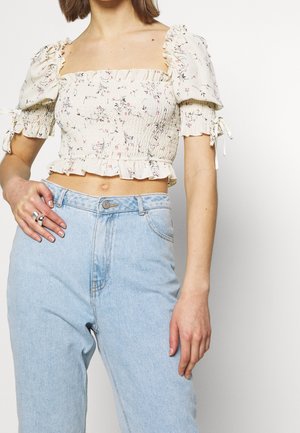 Femme portant un crop top blanc à fleurs avec manches bouffantes et un jean taille haute bleu clair, montrant une main avec des bagues sur le côté gauche.