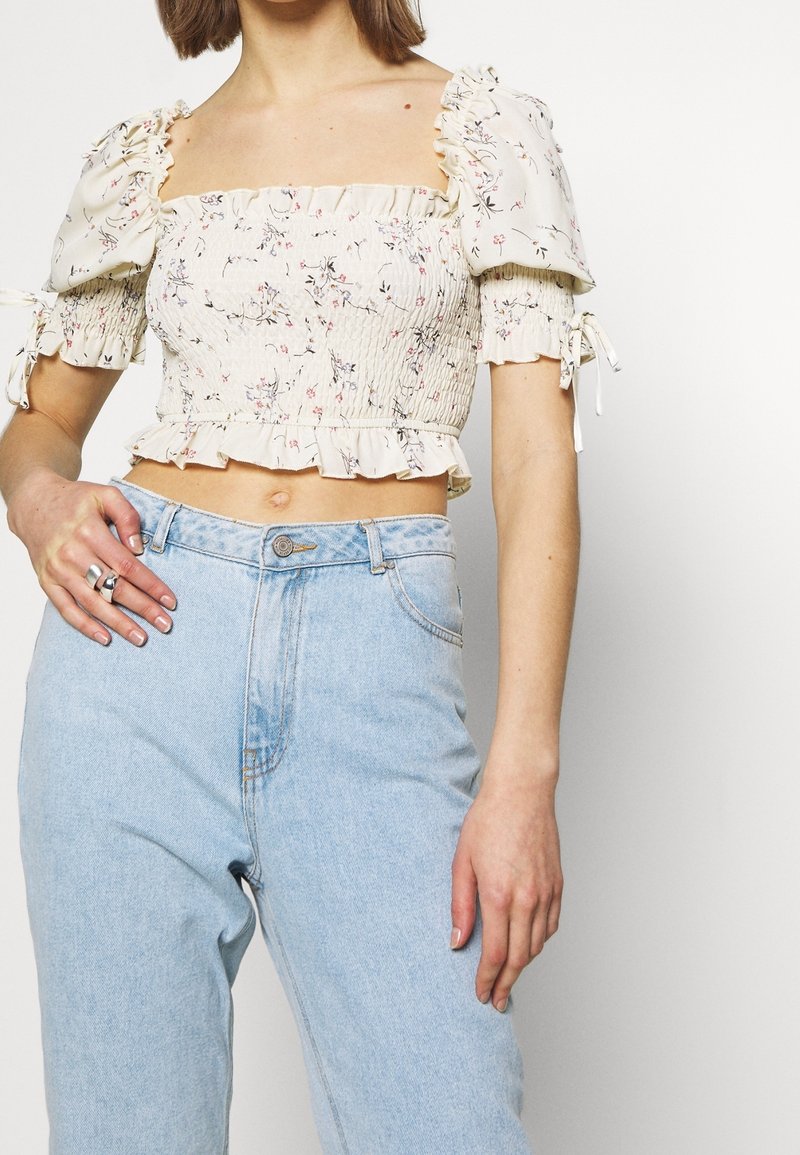 Femme portant un crop top blanc à fleurs avec manches bouffantes et un jean taille haute bleu clair, montrant une main avec des bagues sur le côté gauche.
