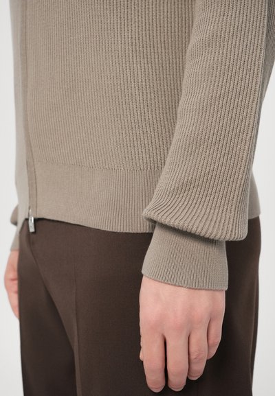 Μπεζ πουλόβερ πλεκτό με ραβδώσεις και κλείσιμο με φερμουάρ, που διαθέτει υφή με ανάγλυφο μοτίβο και εφαρμοστά μανίκια, σε συνδυασμό με σκούρο καφέ παντελόνι.