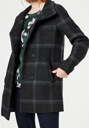 Femme portant un manteau boutonné à carreaux gris foncé par-dessus un top floral vert, avec une main dans la poche du manteau.