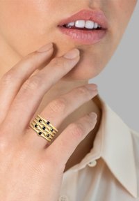 Goldring mit durchbrochenem Design, der mehrere blaue Edelsteine in einem diagonalen Muster zeigt. Glatte Oberfläche, getragen an einer Hand, die das Gesicht berührt.