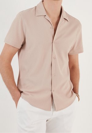 Man wearing short-sleeve, beige ribbed button-up shirt tucked into white pants, with hands in pockets, against plain background.