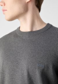 Pilkas megztinis, pagamintas iš minkštos medžiagos, su apvaliu kaklu ir mažuoju išsiuvinėtu "BOSS" logotipu kairėje krūtinės pusėje.