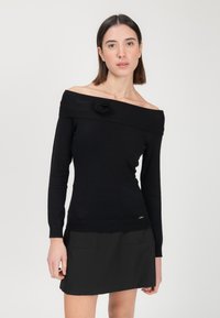 Schwarzer Off-Shoulder-Pullover mit einer strukturierten Rosenakzent; gerippte Bündchen und Saum, weicher Stoff, figurbetonendes Design. Tragen mit einem schwarzen Rock.