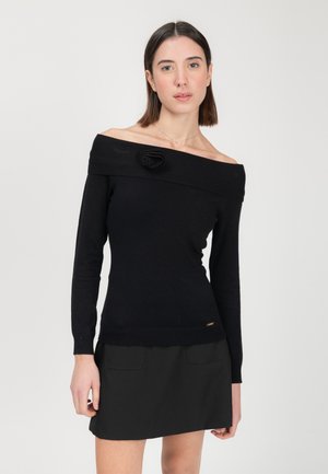 Maglione nero off-the-shoulder con un'accento di rosa testurizzato; polsini e orlo a costine, tessuto morbido, design aderente. Indossato con una gonna nera.