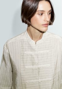 Camicia button-up beige chiaro con righe verticali, colletto a bandana e anelli decorativi. Realizzata in tessuto leggero con una finitura testurizzata.