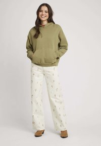 Un sweat à capuche vert olive avec une poche frontale, associé à un pantalon crème à motifs floraux. Des bottines marron à la cheville complètent la tenue.