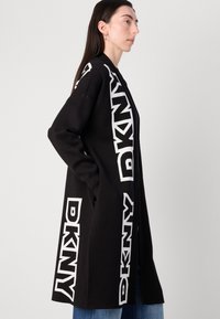 Dolga črna bunda z velikimi belimi DKNY logotipi na straneh, s preprostim, čistim dizajnom in sproščenim krojem.