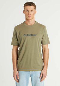 Olive spalvos medvilnės marškinėliai trumpomis rankovėmis, apvaliu iškirpimu ir tamsiai pilku "CHASIN'" logo ant krūtinės. Lygus paviršius, klasikinis siluetas.