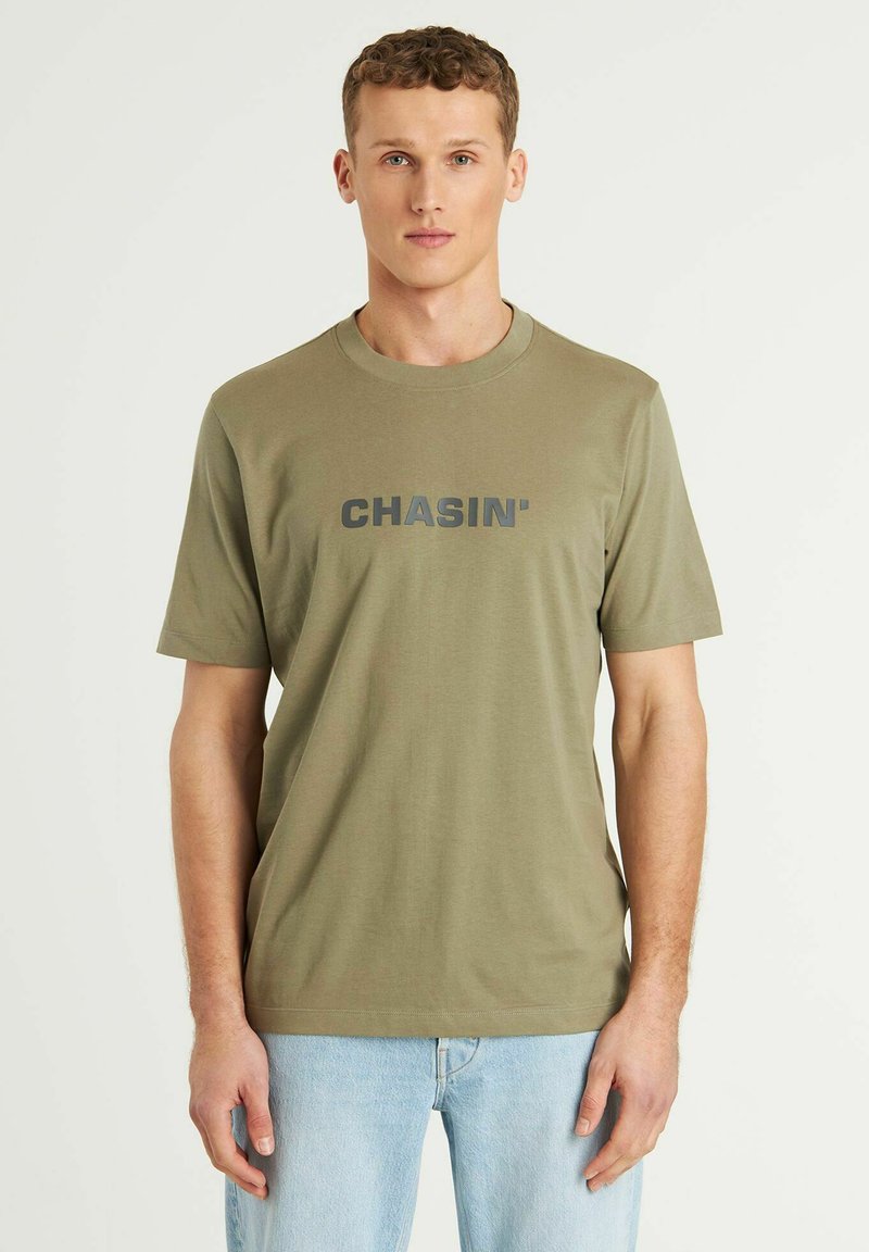 Olive spalvos medvilnės marškinėliai trumpomis rankovėmis, apvaliu iškirpimu ir tamsiai pilku "CHASIN'" logo ant krūtinės. Lygus paviršius, klasikinis siluetas.
