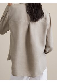 Blouse en lin marron clair à coupe décontractée, col boutonné et manches longues. Caractérisée par un détail plissé au dos et une texture lisse.