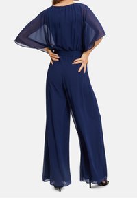 Femme portant un pantalon large bleu marine et un haut transparent assorti avec des manches chauve-souris, debout, les mains sur les hanches, de dos.