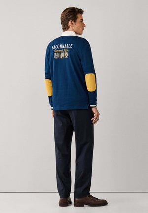 Homme portant une chemise bleu marine à manches longues avec des coudières jaunes et le texte « "Façonnable French Alps" » dans le dos, pantalon foncé et bottes marron, de profil.