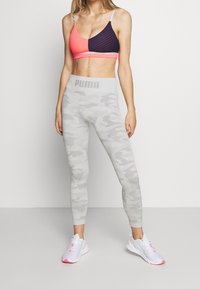 Sportovní podprsenka v růžové a námořnické barvě se pruhovaným vzorem; šedé legíny s maskáčovým vzorem a logem Puma; doplněno o bílé a růžové sportovní boty.