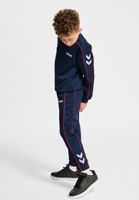 Marinblå träningsoverall med röda accentlinjer, med en logotyp på bröstet och chevron-mönster på byxorna. Matchas med svarta sneakers.