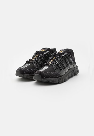 Black athletic shoes with a textured upper, featuring zigzag stitching, reflective accents, and contrasting gold branding on the laces.