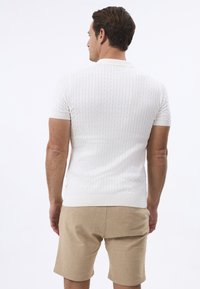 Suéter corto de manga corta de punto blanco con patrón de cable y cuello acanalado, combinado con pantalones cortos beige, mostrando una textura suave.