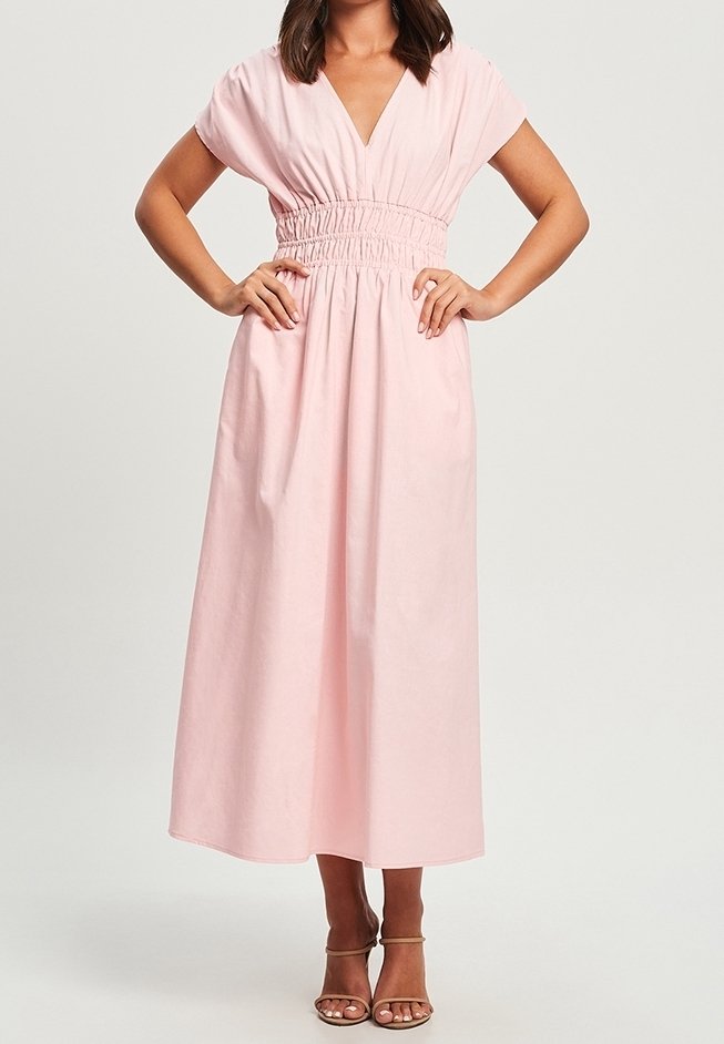 Femme portant une robe légère rose pâle, à manches courtes et mi-longue, avec une taille froncée, debout les mains sur les hanches et portant des sandales à talons beiges.