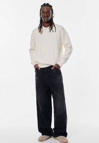 Helle beige Langarm-Henley-Oberteil mit drei Knöpfen, kombiniert mit locker sitzenden dunklen Jeans und beigen Schuhen. Einfaches Design, weicher Stoff.