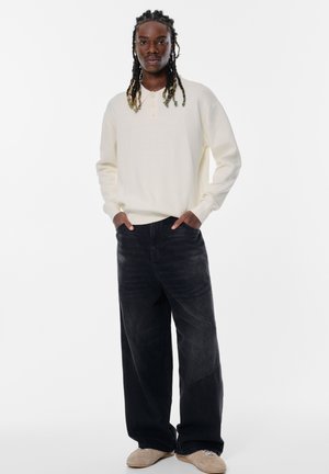 Helle beige Langarm-Henley-Oberteil mit drei Knöpfen, kombiniert mit locker sitzenden dunklen Jeans und beigen Schuhen. Einfaches Design, weicher Stoff.