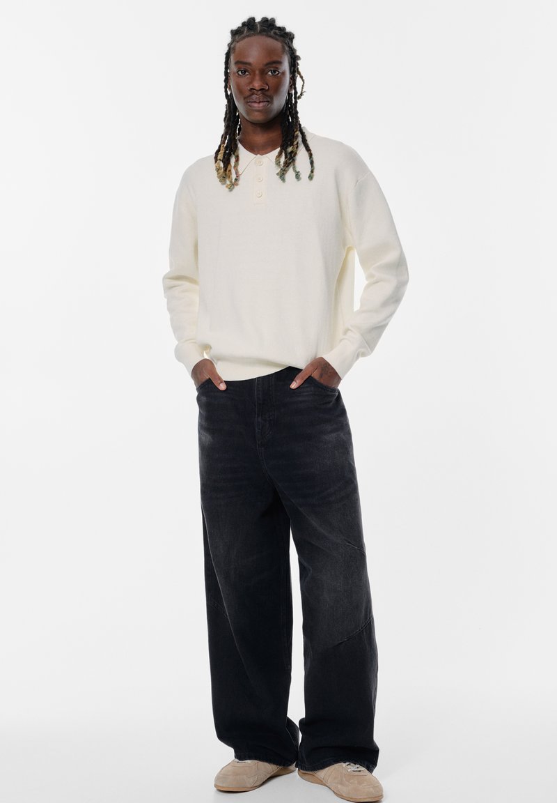 Helle beige Langarm-Henley-Oberteil mit drei Knöpfen, kombiniert mit locker sitzenden dunklen Jeans und beigen Schuhen. Einfaches Design, weicher Stoff.