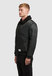 Bomber jacket nero con colletto in finta shearling, tasche con zip sul petto e sulle maniche, e polsini a costine. Tessuto liscio e imbottito.