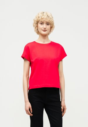 Mujer con cabello rubio corto y rizado, lleva una blusa de manga corta de color rojo brillante y pantalones negros, de pie contra un fondo claro y liso.