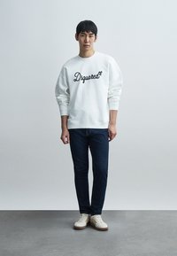 Νεαρός άνδρας που στέκεται, φοράει λευκό φούτερ με το λογότυπο "Dsquared²", σκουρόχρωμα μπλε τζιν και λευκά αθλητικά παπούτσια, μπροστά από απλό γκρι φόντο.