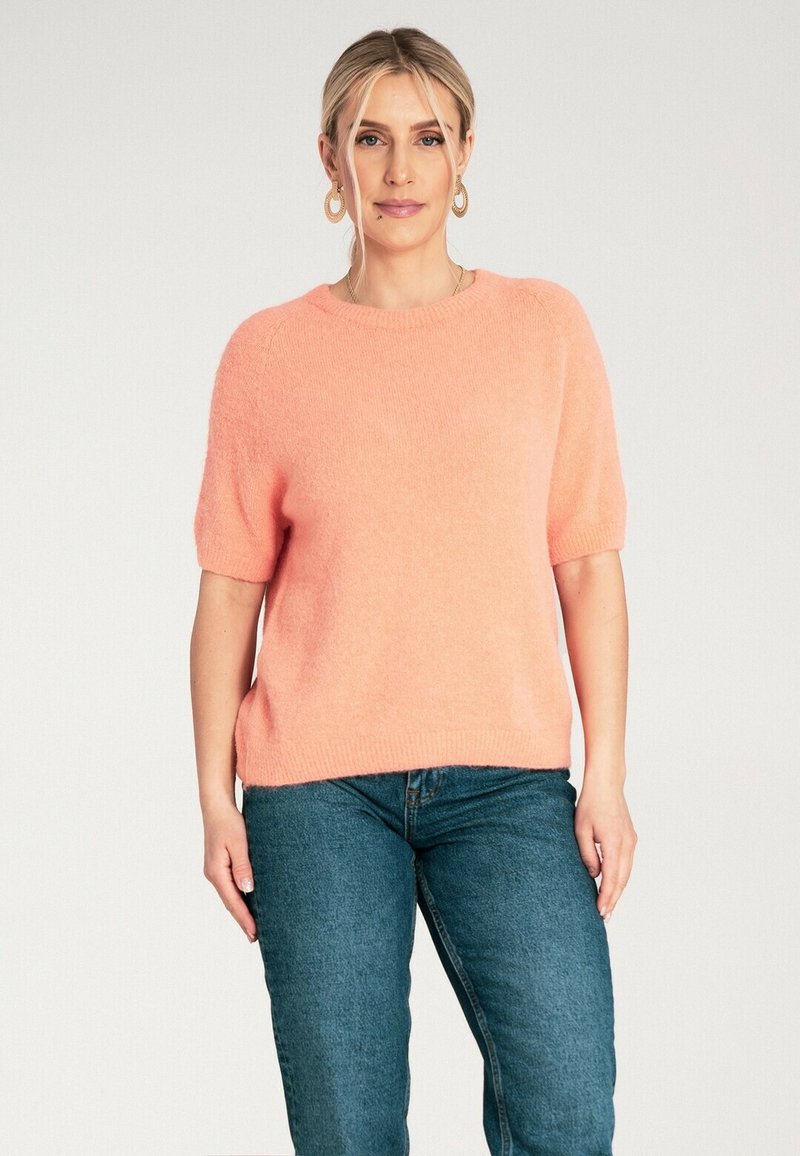 Krótki rękaw, lekki sweter z miękkiego, jasnoróżowego materiału. Posiada okrągły dekolt i luźny krój, zestawiony z niebieskimi dżinsami.