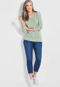 Groene en blauwe textuur gebreide top met driekwart mouwen, gecombineerd met denim enkelbroeken en witte sneakers. Lichte achtergrond.