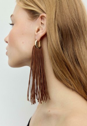 Profil af en kvinde med langt lysebrunt hår, der bærer en stor guldfarvet og brun kvastørering, som går ned over hendes skulder.