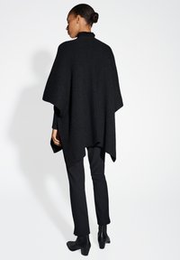 Zwarte poncho met een gestructureerd breisel en een gerimpelde hoge kraag, met een losse pasvorm en subtiele glinstering, gecombineerd met zwarte broek en laarzen met hak.