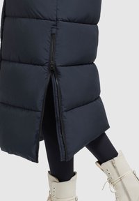 Marinblå pufferjacka med dragkedja i sidan, quiltad struktur och hög krage. I kombination med vita snörstövlar och svarta leggings.