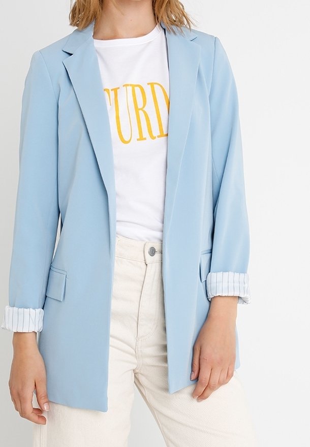 Femme portant un blazer bleu clair avec des poignets rayés retroussés, un t-shirt blanc avec un texte jaune, et un pantalon taille haute écru.