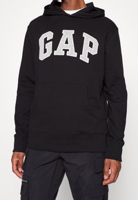 Black hoodie with a kangaroo pocket, featuring a gray "GAP" logo across the chest, made from soft fabric with a relaxed fit and ribbed cuffs.