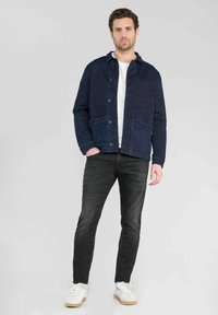Chaqueta de algodón azul marino con cierre de botones, bolsillos delanteros y un diseño casual, combinada con unos vaqueros negros y zapatillas blancas.