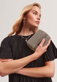 Donna bionda in un abito nero plissettato tiene in mano una borsa argentata con strass, indossando una collana di catena argentata con perle e orecchini a cerchio coordinati.