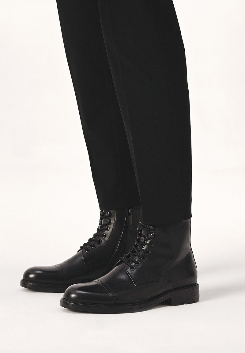 Bocanci negri din piele, cu vârful rotund, șireturi în față și fermoar pe lateral. Design minimalist cu o textură netedă și o talpă robustă.