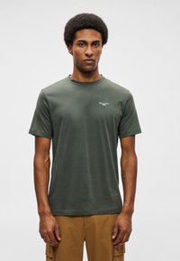 Zaļš kokvilnas T-krekls ar apaļu kaklu, īsām piedurknēm un mazu baltu logo kreisajā krūtī. Gluda tekstūra ar brīvu piegriezumu.